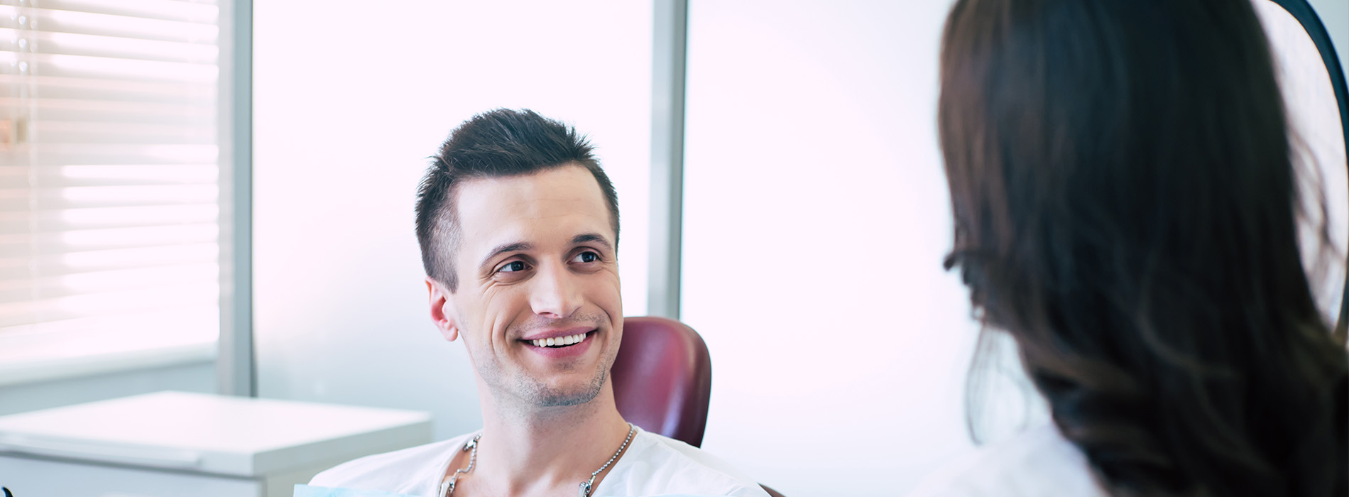 The image shows a man sitting in a dental chair, receiving treatment from a dentist who is standing behind him.