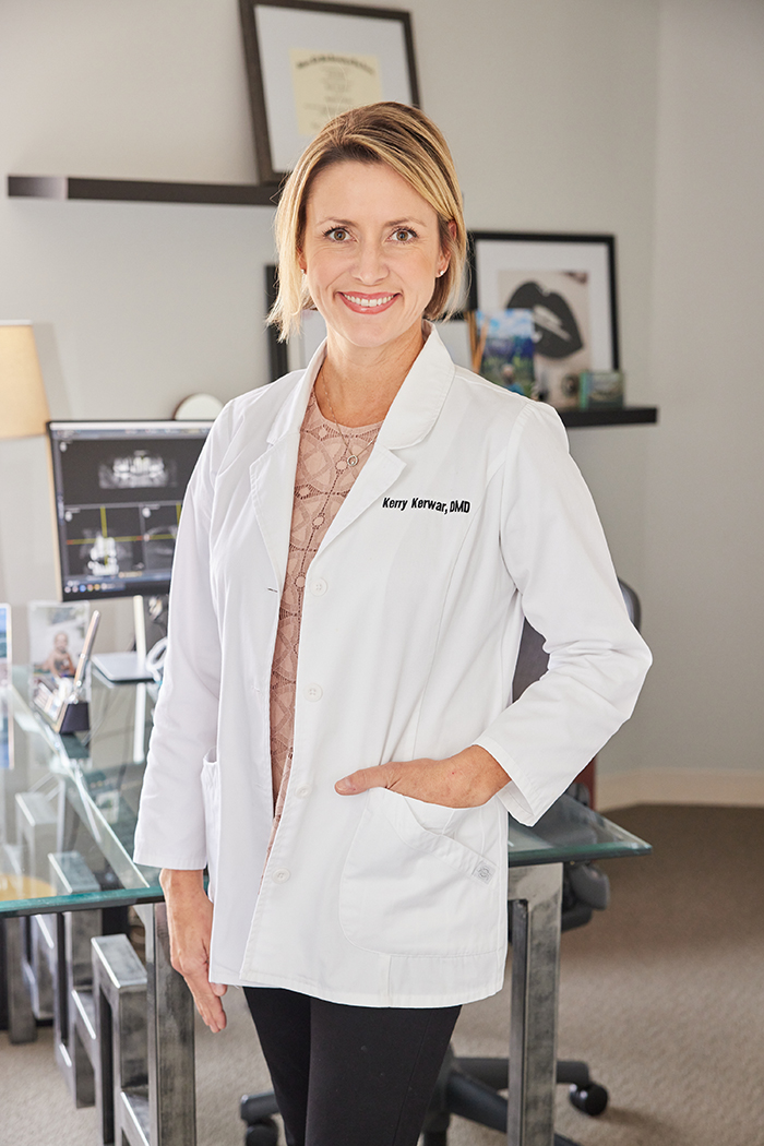 A woman wearing a white lab coat stands confidently in an office setting, posing for a portrait with a smile.